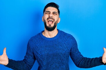 Handsome man with beard wearing casual winter sweater crazy and mad shouting and yelling with aggressive expression and arms raised. frustration concept.