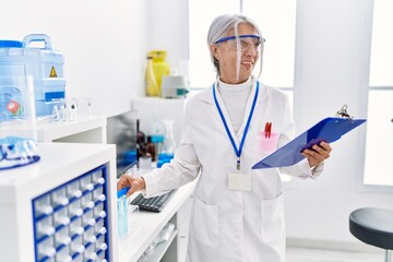 Middle age grey-haired woman wearing scientist uniform holding clipboard and test tube at laboratory