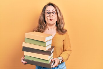 Middle age caucasian woman holding a pile of books making fish face with mouth and squinting eyes, crazy and comical.
