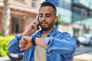 Young hispanic man talking on the smartphone looking watch at street