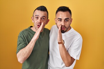 Homosexual couple standing over yellow background hand on mouth telling secret rumor, whispering...
