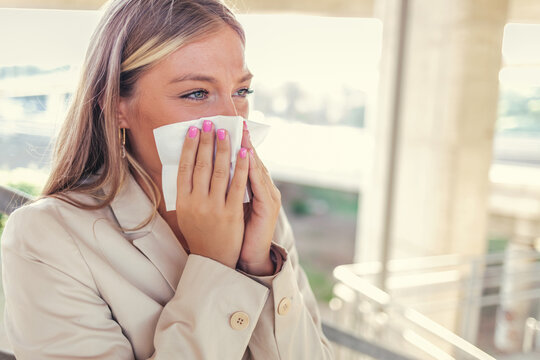 Portrait Of An Uncomfortable Looking Young Woman Blowing Her Nose With A Tissue Outside Of A Building During The Day. Woman Uses Tissue Paper Sneezing Due To Having Pollen Allergy.