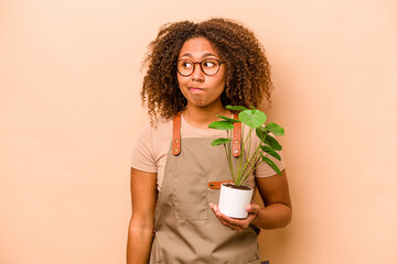 Young gardener African American woman holding plant isolated on beige background confused, feels...