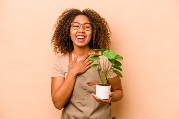 Young gardener African American woman holding plant isolated on beige background laughs out loudly...