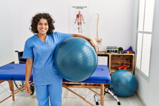 Young hispanic woman wearing physio therapist uniform holding fit ball at clinic