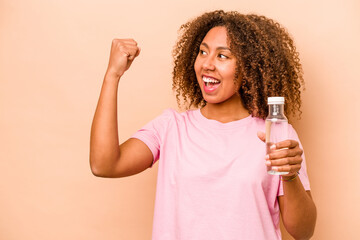 Young African American woman holding a bottle of water isolated on beige background raising fist...