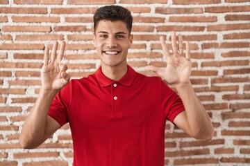 Young hispanic man standing over bricks wall showing and pointing up with fingers number eight while smiling confident and happy.