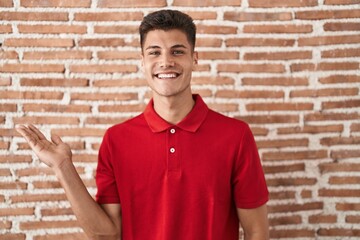 Young hispanic man standing over bricks wall smiling cheerful presenting and pointing with palm of hand looking at the camera.