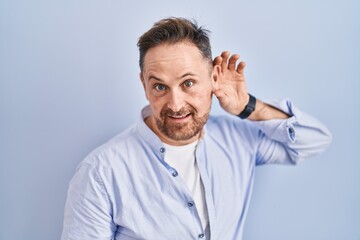 Obraz premium Middle age caucasian man standing over blue background smiling with hand over ear listening an hearing to rumor or gossip. deafness concept.