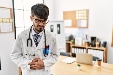 Hispanic man with beard wearing doctor uniform and stethoscope at the office with hand on stomach because nausea, painful disease feeling unwell. ache concept.