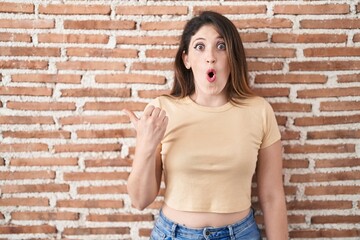 Young brunette woman standing over bricks wall surprised pointing with hand finger to the side, open mouth amazed expression.