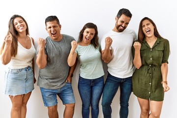 Group of young hispanic friends standing together over isolated background very happy and excited doing winner gesture with arms raised, smiling and screaming for success. celebration concept.