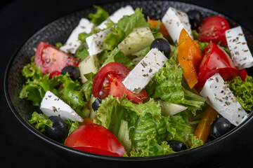 Healthy greek salad of fresh vegetables with tomatoes, lettuce, bell pepper, cucumbers, olives, feta cheese in plate on black stone background. Healthy food, vegetarian dieting, close up