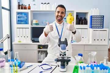 Young hispanic man with beard working at scientist laboratory success sign doing positive gesture with hand, thumbs up smiling and happy. cheerful expression and winner gesture.