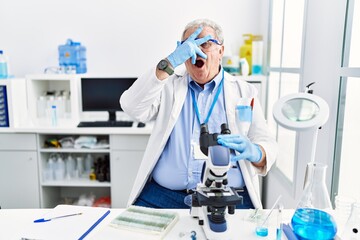 Fototapeta premium Senior caucasian man working at scientist laboratory peeking in shock covering face and eyes with hand, looking through fingers with embarrassed expression.