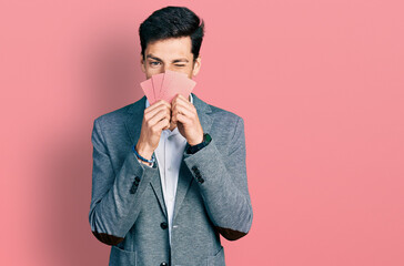 Young hispanic business man covering face with cards winking looking at the camera with sexy...