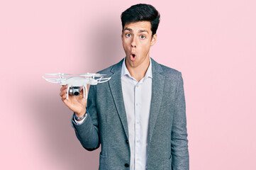 Young hispanic business man holding drone scared and amazed with open mouth for surprise, disbelief...