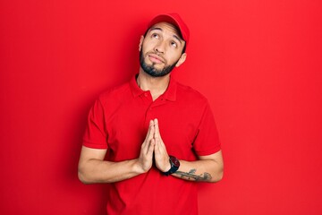 Hispanic man with beard wearing delivery uniform and cap begging and praying with hands together with hope expression on face very emotional and worried. begging.