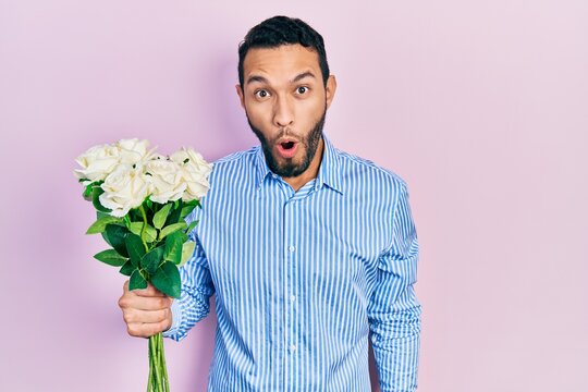 Hispanic man with beard holding bouquet of white flowers scared and amazed with open mouth for surprise, disbelief face