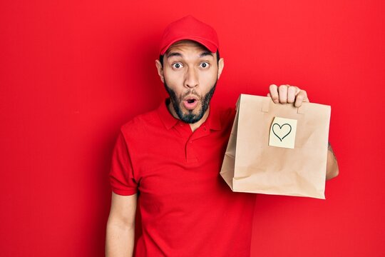 Hispanic man with beard holding take away paper bag with heart reminder scared and amazed with open mouth for surprise, disbelief face