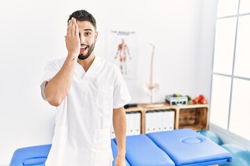 Obraz premium Young handsome man with beard working at pain recovery clinic covering one eye with hand, confident smile on face and surprise emotion.