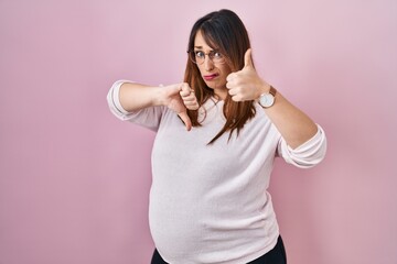 Pregnant woman standing over pink background doing thumbs up and down, disagreement and agreement expression. crazy conflict