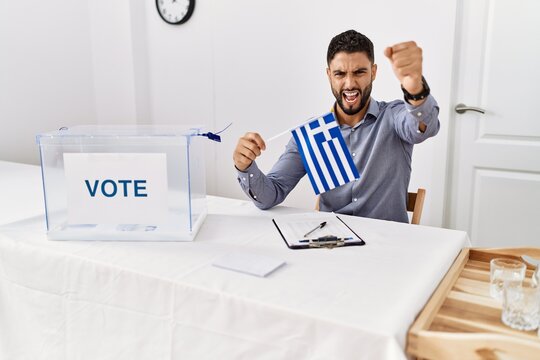 Young handsome man with beard at political campaign election holding greece flag annoyed and frustrated shouting with anger, yelling crazy with anger and hand raised