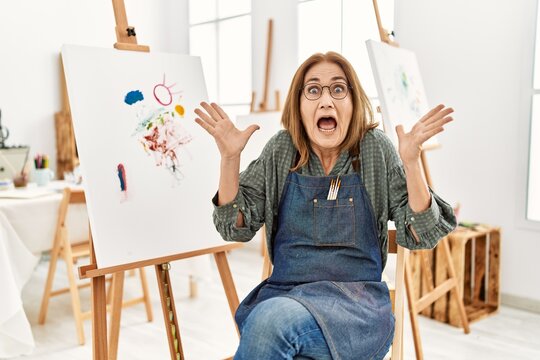 Middle age artist woman at art studio celebrating crazy and amazed for success with arms raised and open eyes screaming excited. winner concept