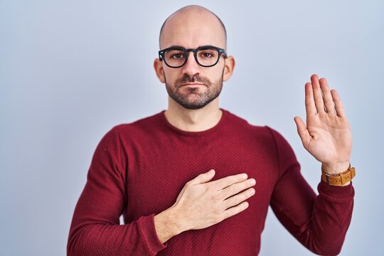 Young bald man with beard standing over white background wearing glasses swearing with hand on chest and open palm, making a loyalty promise oath