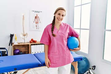 Young caucasian woman wearing physiotherapist uniform holding fit ball at physiotherapy clinic