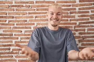 Young caucasian man standing over bricks wall smiling cheerful offering hands giving assistance and acceptance.