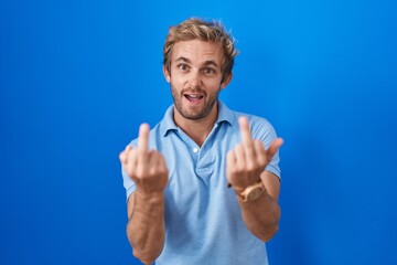 Caucasian man standing over blue background showing middle finger doing fuck you bad expression,...