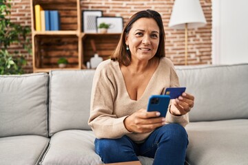Middle age hispanic woman using smartphone and credit card sitting on sofa at home