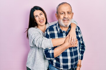 Naklejka premium Young brunette woman and senior man standing over pink background. Daughter and father hugging and bonding together as happy family