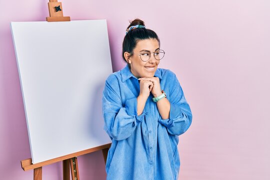 Young hispanic woman standing by painter easel stand laughing nervous and excited with hands on chin looking to the side