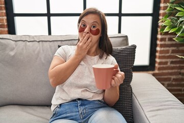 Hispanic girl with down syndrome sitting on the sofa drinking a coffee at home covering mouth with hand, shocked and afraid for mistake. surprised expression