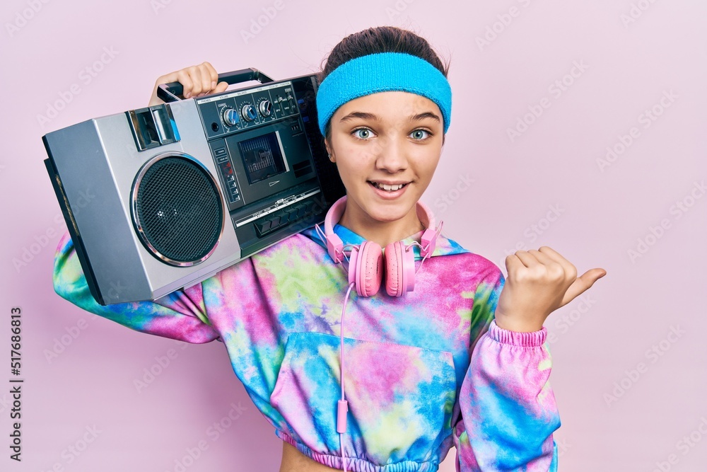 Wall mural young brunette girl wearing workout clothes and holding boombox pointing thumb up to the side smilin