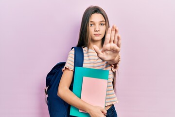 Young brunette girl holding student backpack and books with open hand doing stop sign with serious and confident expression, defense gesture