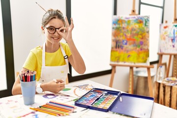 Young brunette teenager at art studio doing ok gesture with hand smiling, eye looking through...