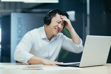 Asian businessman watching sports match in office, man looking disappointed in laptop, using headphones to listen