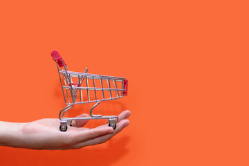 Small shopping trolley in hand on an orange background close-up. The concept of buying and selling....