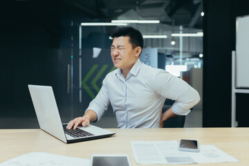 Asian businessman working in office, having severe back pain, overtired worker working with laptop.