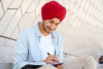 Focused Indian student in traditional turban sitting outdoors with notebook and taking notes,...
