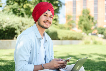 Smiling Indian male student in national turban sitting outdoors with laptop, using smartphone, texting online, chatting in social media network, using dating app. Side view