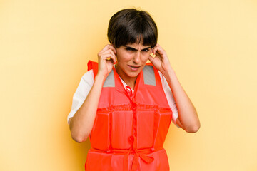 Young caucasian woman wearing life jacket isolated on yellow background covering ears with hands.