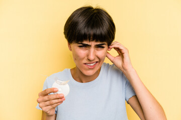 Young caucasian woman holding teeth whitener isolated on yellow background covering ears with hands.