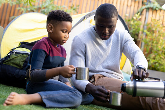Image of smiling african american boy with father drinking tea on the camping