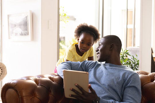 Image of happy african american father and daughter sitting on sofa with tablet