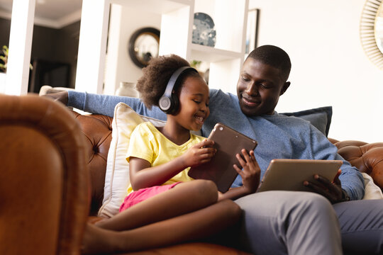 Image of happy african american father and daughter sitting on sofa with tablets