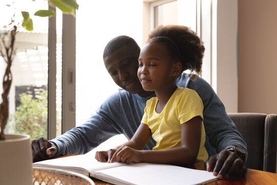 Image of happy african american father and daughter with sight disability reading in braille